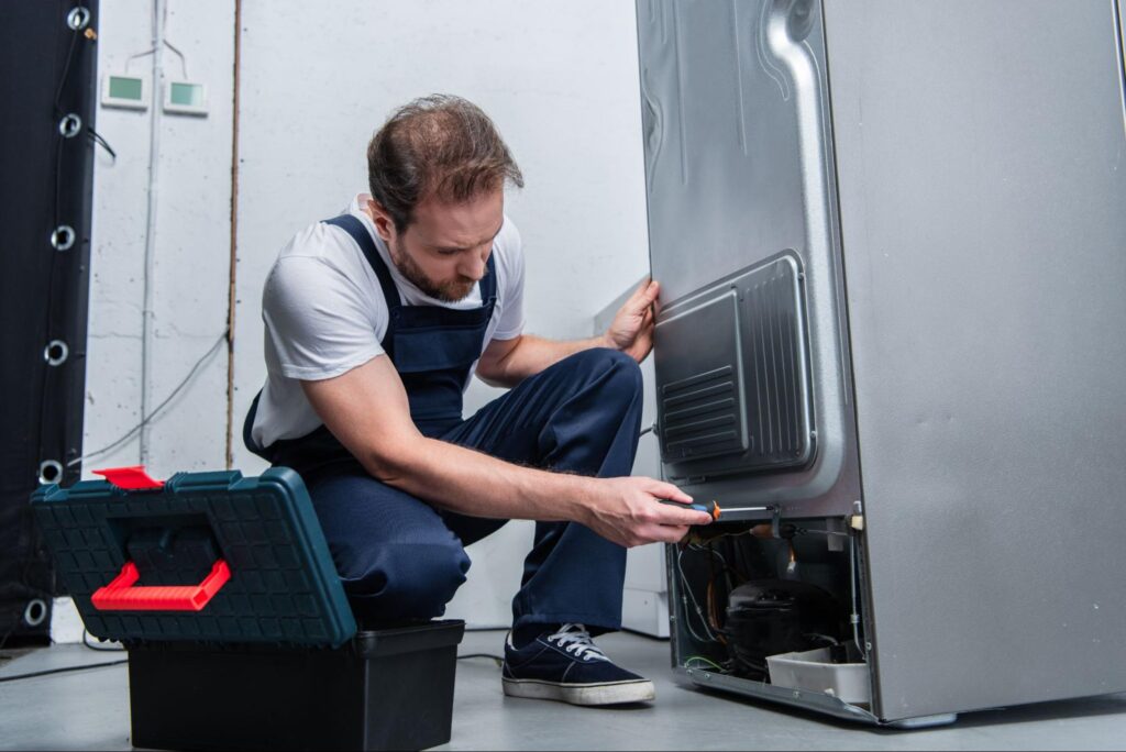 Man in overalls doing repairs on a refrigerator.