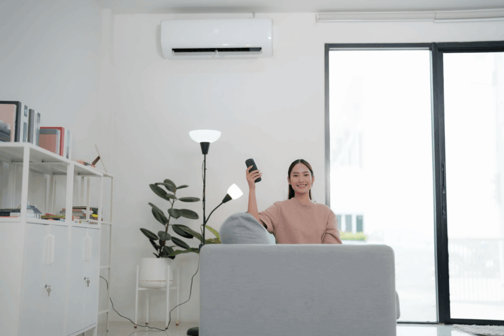 A woman sitting on a couch adjusts a ductless mini split behind her with a remote control.
